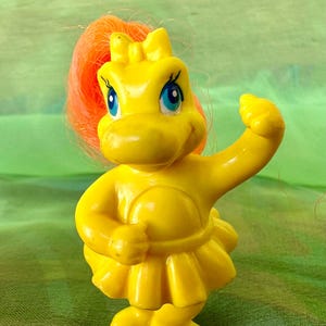 May include: A yellow plastic toy figure of a hippopotamus-like creature with blue eyes and orange hair. The figure is wearing a yellow dress and has one arm raised. The toy is a vintage collectible.