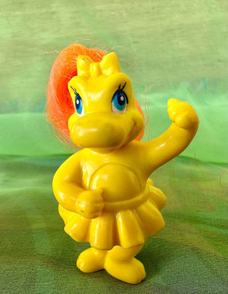 May include: A yellow plastic toy figure of a hippopotamus-like creature with blue eyes and orange hair. The figure is wearing a yellow dress and has one arm raised. The toy is a vintage collectible.