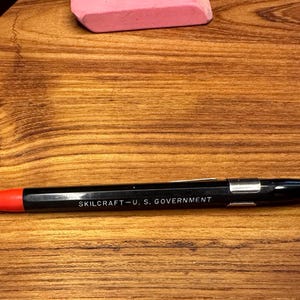 May include: Black and silver Skilcraft pen with a red tip, featuring the text "SKILCRAFT - U.S. GOVERNMENT." The pen is lying on a wooden surface, alongside a pink eraser and a calculator.