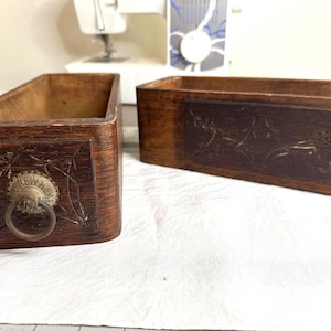 Könnte beinhalten: Zwei rechteckige Holzschubladen mit dunkelbraunem Finish. Die Frontplatten weisen geschnitzte Blumenmuster auf. Eine Schublade enthält einen runden Messinggriff mit der Prägung NEW HOME auf der Metallplatte um den Ring herum.