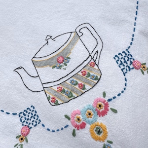 May include: A white fabric cloth featuring hand-embroidered designs. The central motif is a teapot outlined in black thread with grey stripes and small floral accents. Surrounding the teapot are blue stitched lines and clusters of pink, yellow, and blue flowers.