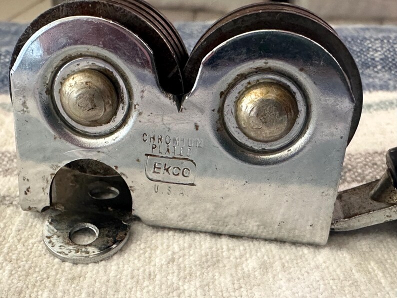 Puede incluir: Primer plano de un afilador de cuchillos cromado vintage. El afilador tiene dos ruedas giratorias y un mango. El texto "CHROMIUM PLATED" y "Ekco U.S.A." son visibles en el cuerpo met&aacute;lico.