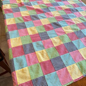 May include: A patchwork quilt featuring a grid of square fabric patches in a gingham pattern. The squares alternate in colors including light blue, pink, yellow, green, red, and navy blue, creating a multi-colored checkered textile design.