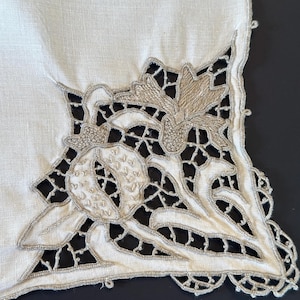 May include: Close-up of a cream linen textile with an embroidered floral design. The embroidery uses silver-coloured thread to outline floral shapes and create a delicate, openwork pattern. The textile's edge has a scalloped trim.