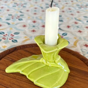 May include: A glossy lime green ceramic candle holder shaped like a large leaf with a central socket holding a single off-white taper candle with a charred black wick. The base of the holder features raised veins and a smooth, reflective finish.