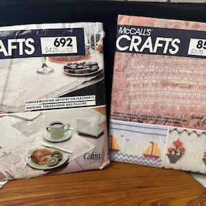 Puede incluir: Dos folletos de patrones McCall's Crafts vintage. Uno presenta un diseño de mantel individual y camino de mesa, con el texto "Candlewicking Artistry on Placemats, Napkins, Tablerunner and Pillows". El otro presenta bordados y bordes bordados.