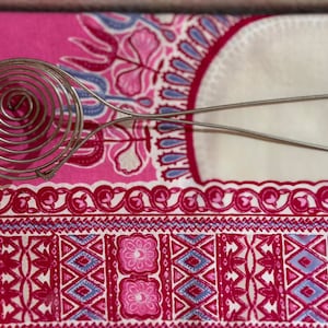 May include: A stainless steel spiral whisk with a long handle, resting on a pink and white patterned fabric. The whisk's spiral head is designed for efficient mixing. The fabric features floral and geometric designs.