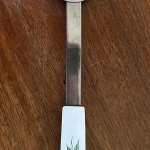 Op de afbeelding: Een vintage serveervork met een roestvrijstalen kop en een witte handgreep. De handgreep is versierd met roze bloemen en groene bladeren. De vork is ontworpen voor het serveren van voedsel.