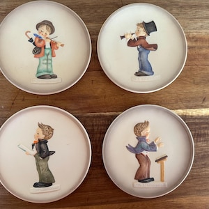 May include: Set of four decorative plates featuring children playing musical instruments. Each plate has a raised relief design of a child playing a violin, flute, singing, and conducting. The plates are white with a light pink border.