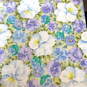 May include: A fabric featuring a dense floral pattern of pansies in shades of white, light blue, lavender, and purple. The flowers are set against a light green background with visible green foliage, creating a continuous botanical print.
