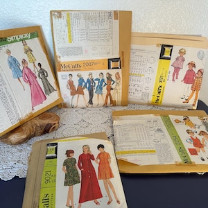 May include: A collection of vintage sewing patterns from brands like Simplicity and McCall's. The patterns feature illustrations of dresses and coats in various colors. The patterns are in tan envelopes with yellow and pink labels.
