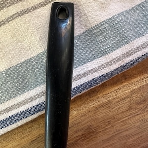 Puede incluir: Primer plano de un mango negro de un utensilio de cocina, posiblemente un abrelatas. El mango es liso y tiene un agujero en la parte superior. Est&aacute; unido a una base de metal plateado. El fondo incluye un pa&ntilde;o a rayas y una superficie de madera.