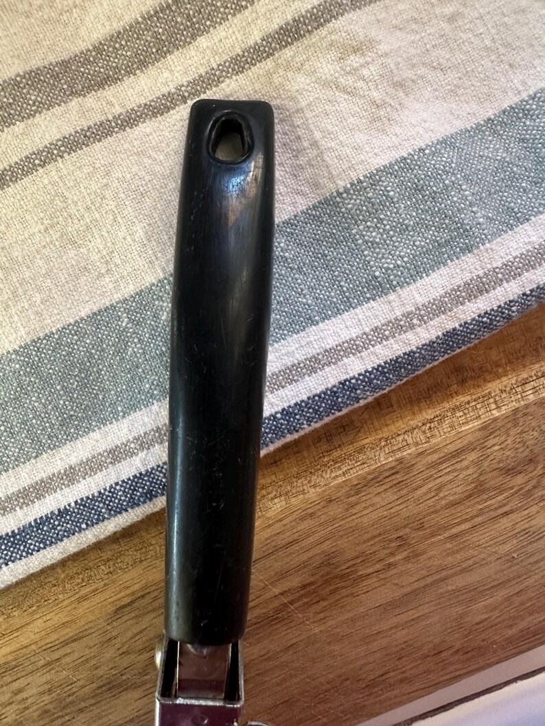 Puede incluir: Primer plano de un mango negro de un utensilio de cocina, posiblemente un abrelatas. El mango es liso y tiene un agujero en la parte superior. Est&aacute; unido a una base de metal plateado. El fondo incluye un pa&ntilde;o a rayas y una superficie de madera.