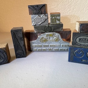 Puede incluir: Una colección de bloques de impresión vintage de madera y metal. Los bloques presentan varios diseños, incluyendo un coche, un pájaro, un logotipo y texto. Los bloques son de varios tonos de marrón, negro y plateado.