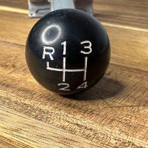 Könnte beinhalten: Schwarzer Schaltknauf mit weißen Markierungen für Rückwärtsgang (R), ersten (1), zweiten (2), dritten (3) und vierten (4) Gang. Der Knauf ist kugelförmig und liegt auf einer Holzoberfläche.