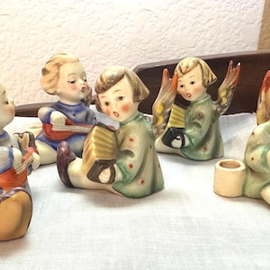 May include: Five porcelain figurines of angels playing musical instruments. The angels are dressed in colorful robes and have wings. They are holding a guitar, accordion, and a small trumpet.