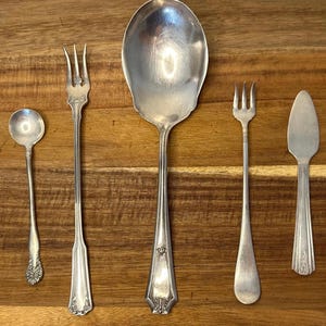 Puede incluir: Colección de cubiertos antiguos de plata, que incluye una cuchara grande para servir, dos tenedores, una cuchara pequeña y un cuchillo de mantequilla. La cubertería es de metal brillante color plata y está dispuesta sobre una superficie de madera.