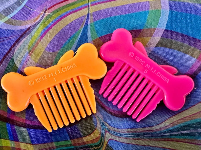 May include: Two plastic combs shaped like bones, one orange and one pink, with comb teeth. The combs have a fish-like design and the text "1992 M.I.I. CHINA" printed on them. The orange comb has the number "1" and the pink comb has the number "5".