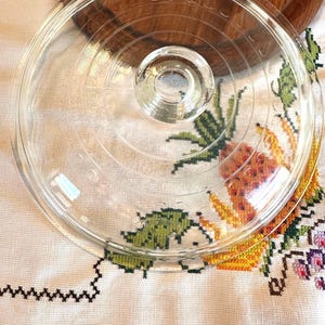 Puede incluir: Una tapa de vidrio transparente con un asa circular. La tapa descansa sobre una superficie de madera y un mantel blanco con bordados de punto de cruz coloridos. La tapa tiene anillos concéntricos grabados en el vidrio.