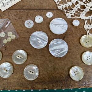 May include: A collection of round, iridescent mother-of-pearl buttons in various sizes. Some buttons feature two holes, while others have four holes. Several smaller buttons are contained within a clear plastic bag, and others are loose.