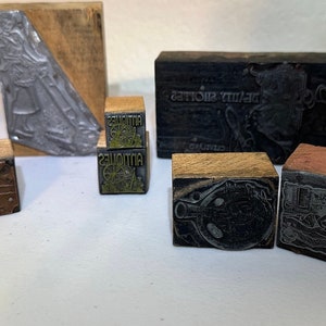 May include: A collection of vintage wood and metal printing blocks. The blocks feature various designs, including a woman in a dress, a man with a hat, and a geometric pattern. The blocks are in various shades of brown, black, and silver.
