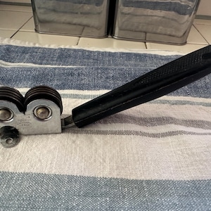 Puede incluir: Un afilador de cuchillos manual con un mango negro y dos juegos de muelas. El cuerpo met&aacute;lico tiene un acabado plateado. El afilador est&aacute; dise&ntilde;ado para uso en la cocina y descansa sobre un pa&ntilde;o a rayas azules y blancas.
