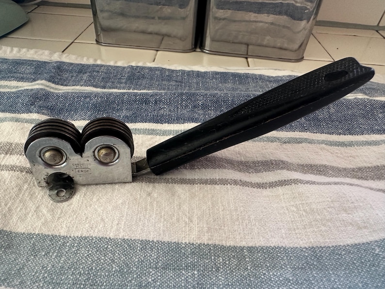 Puede incluir: Un afilador de cuchillos manual con un mango negro y dos juegos de muelas. El cuerpo met&aacute;lico tiene un acabado plateado. El afilador est&aacute; dise&ntilde;ado para uso en la cocina y descansa sobre un pa&ntilde;o a rayas azules y blancas.