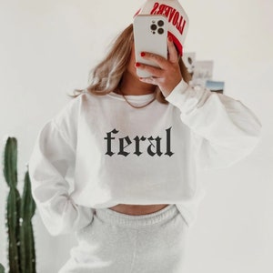 May include: White cropped sweatshirt with the word "feral" printed in black gothic font. The model is wearing a red and white baseball cap.