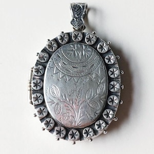 May include: An oval silver locket pendant featuring an engraved floral motif and a crescent pattern on the center plate. The border is decorated with a series of small circular discs, each embossed with a six-pointed star design, and a bail.