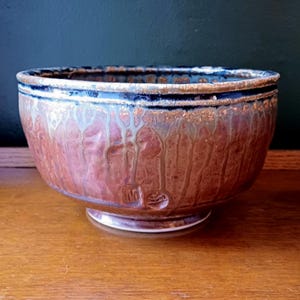 May include: A handcrafted ceramic bowl with a wide, rounded form. The bowl has a reddish-brown glaze with dripped patterns and a blue rim. A small, stamped design is visible near the base. The bowl rests on a small, circular foot.