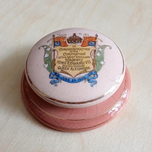 May include: A pink ceramic trinket box with a lid commemorating the coronation of King Edward VII and Queen Alexandra on June 26, 1902. The box has a gold trim and a decorative design.