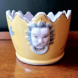 May include: A yellow ceramic bowl with a white, scalloped rim and a white face with a beard. The bowl features a lion's face on the front and a blue floral design on the side. It is set on a wooden surface.