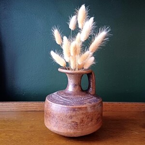 May include: A brown ceramic vase with a handle, filled with fluffy, beige dried flowers. The vase has a textured surface and a decorative band around the neck. The background is a dark green wall, and the vase sits on a wooden surface.