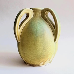 May include: A ceramic vase with a bulbous body and two arched handles extending from the neck to the shoulder. The surface features a mottled glaze in shades of pale yellow and soft green, creating a textured appearance across the entire exterior.