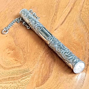 May include: A silver-coloured, cylindrical object with ornate floral and scrollwork detailing. A small chain is attached to one end. The object appears to be a container or holder, possibly for a small item.