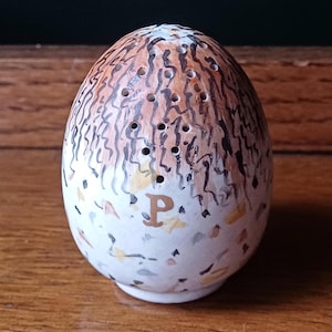 May include: An egg-shaped ceramic salt shaker with a speckled white base and a brown and black patterned top. The shaker has several small holes at the top and a gold-coloured letter "P" near the base. The design is hand-painted.