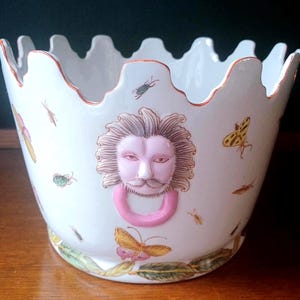 May include: White ceramic planter with a scalloped rim and a lion's head design. The planter features a pink ring, and is decorated with insects and butterflies in shades of yellow, pink, and green. The rim has a gold trim.