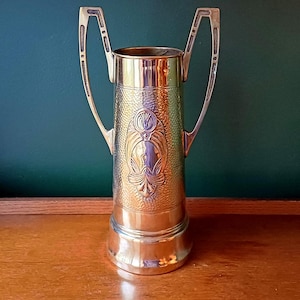 May include: A brass-coloured vase with two curved handles. The vase has a textured, hammered surface and a decorative embossed design in the centre. The vase is on a wooden surface.