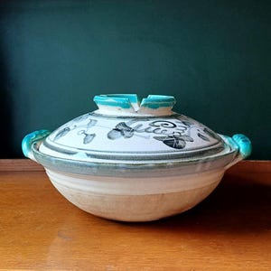May include: A round ceramic lidded casserole dish with a cream-coloured base and a lid featuring black hand-painted leaf and vine motifs. The lid handle and two side handles are glazed in a vibrant turquoise blue, with a notched lid knob.