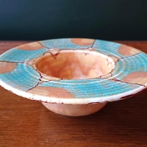 May include: A handcrafted ceramic bowl with a wide, turquoise rim featuring brown and orange accents. The bowl's interior is a warm peach colour, and the exterior has a cream-coloured base. The bowl is a decorative piece.