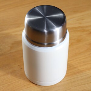 May include: A white insulated food container with a stainless steel lid. The container has a cylindrical shape and a smooth, matte finish. The lid is a brushed metal with a circular design.