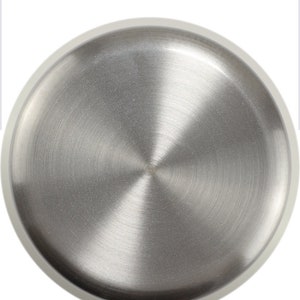 May include: A circular, brushed stainless steel object with a diameter of 3.9 inches. The metal surface has a radial grain pattern, and a white border surrounds the edge. The object is likely a lid or a small plate.