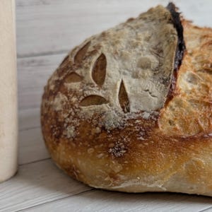 Puede incluir: Una hogaza de pan artesanal con corteza dorada y cortes en forma de hojas. El pan está junto a un rodillo de madera de color claro sobre una superficie de madera blanca. La textura del pan es áspera y atractiva.