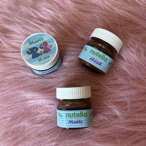 Peut inclure: Trois petits pots en verre remplis de pâte à tartiner au chocolat et aux noisettes. Les pots sont étiquetés avec le texte "Nutella" et le nom "Maëly". Un pot a un personnage de dessin animé bleu et rose sur l'étiquette.