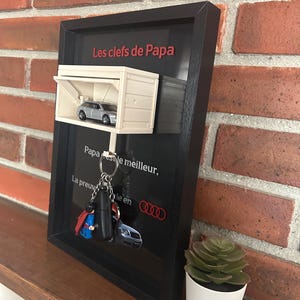Könnte beinhalten: Ein gerahmter Schlüsselhalter mit schwarzem Rahmen und einem weißen Garagenmodell. Die Garage enthält ein silbernes Spielzeugauto. Der Text "Les clefs de Papa" ist rot. Schlüsselanhänger hängen darunter.