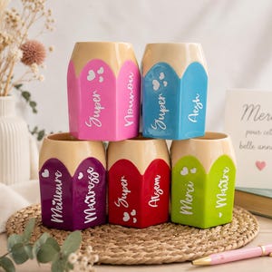Puede incluir: Cinco portalápices de cerámica con forma de lápiz en rosa, azul, morado, rojo y verde lima. Cada uno presenta texto en blanco que incluye 'Super Nounou', 'Super Aesh', 'Meilleure Maitresse', 'Super Atsem' y 'Merci Maitre' con iconos de corazones.
