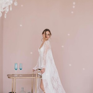 May include: A woman in a white lace robe stands next to a gold bar cart with blue glasses and silver disco balls. The background is a light pink wall.