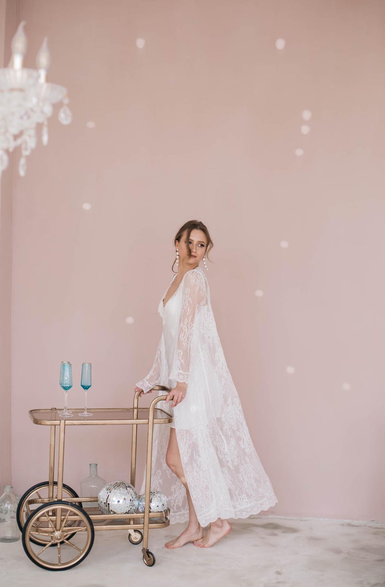 May include: A woman in a white lace robe stands next to a gold bar cart with blue glasses and silver disco balls. The background is a light pink wall.