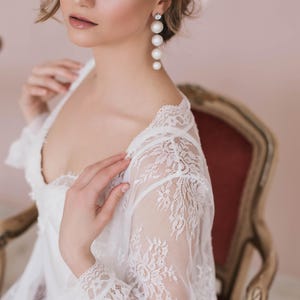 May include: A woman wearing a white lace robe with a deep V-neckline. She is wearing pearl drop earrings.