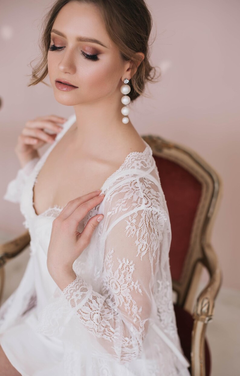 May include: A woman wearing a white lace robe with a deep V-neckline. She is wearing pearl drop earrings.